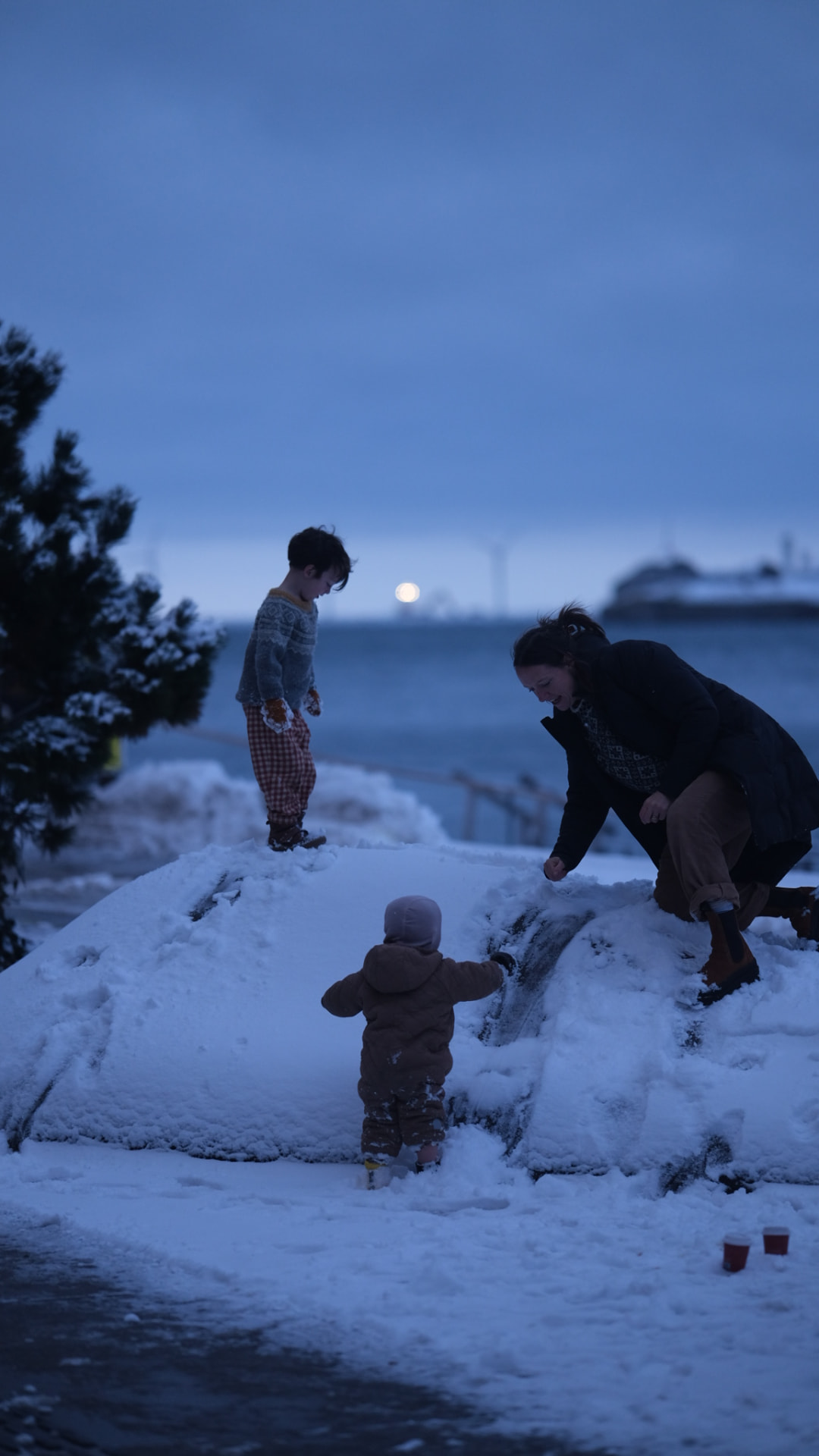 A mother and two children playing on a snowy mound by the sea at blue hour