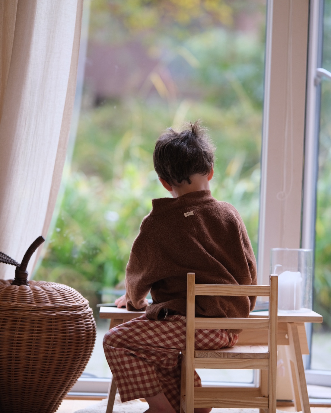 A child in a brown fleece sitting at a small wooden table by a garden window